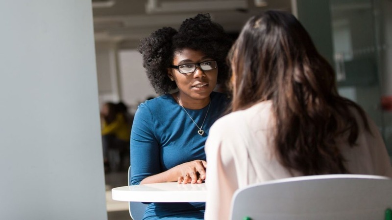Two people talking at a round table in an indoor setting, one facing the camera in a blue top.